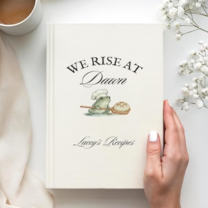 May include: A cream-colored hardcover book with the text "WE RISE AT Dawn" and "Lacey's Recipes" printed in black. The cover features a small illustration of a green frog wearing a white chef hat and holding a wooden peel with a loaf of bread.