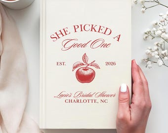 May include: A cream-colored hardcover book featuring red text that reads "SHE PICKED A Good One", "EST. 2026", "Lacie's Bridal Shower", and "CHARLOTTE, NC". A red illustration of an apple with leaves is centered between the text elements.