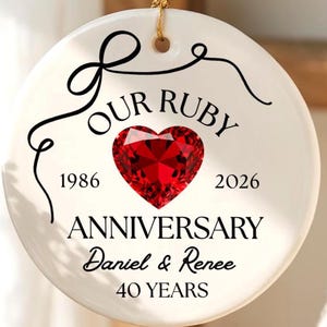 Peut inclure: Une décoration ronde en céramique blanche suspendue par un cordon doré. Elle présente un motif de cœur rouge, le texte "OUR RUBY ANNIVERSARY", les années "1986" et "2026", les noms "Daniel & Renee" et "40 YEARS" avec une volute noire.