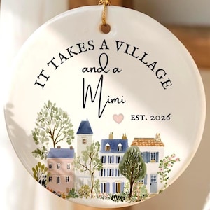 Könnte beinhalten: Ein rundes, weißes Keramikornament, das an einer goldenen Schnur hängt. Der Text lautet IT TAKES A VILLAGE and a Mimi EST. 2026 mit einem kleinen rosa Herz. Darunter befindet sich eine Aquarellillustration von Häusern und Bäumen in Blau und Grün.