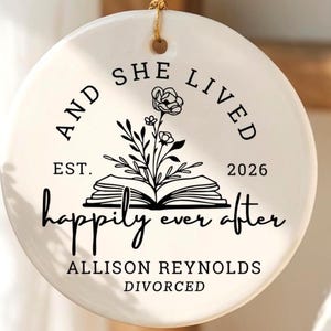 May include: A round white ceramic ornament hanging by a gold string. The front features black text reading AND SHE LIVED, EST. 2026, happily ever after, ALLISON REYNOLDS, and DIVORCED, with a central graphic of an open book and a flower.