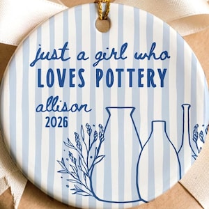 Peut inclure: Une décoration en céramique ronde avec un motif à rayures verticales bleu clair et blanc. Le texte "just a girl who LOVES POTTERY", "allison" et "2026" est inscrit en bleu. Un dessin au trait bleu de poterie et de feuillage est visible.