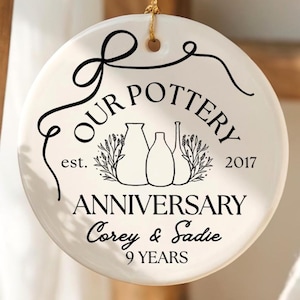 May include: A round white ceramic ornament hanging by a gold string. The front features black text reading OUR POTTERY est. 2017 ANNIVERSARY Corey & Sadie 9 YEARS, with a line drawing of three vases and decorative flourishes.
