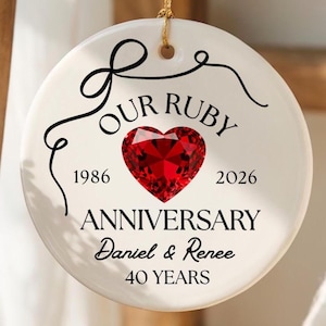 Puede incluir: Un adorno redondo de cerámica blanca colgado de un cordón dorado. El frente presenta un gráfico de corazón rojo, el texto "OUR RUBY ANNIVERSARY", los años "1986" y "2026", los nombres "Daniel & Renee" y el texto "40 YEARS" en letras negras.