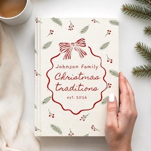 May include: A hardcover book with a cream-colored cover featuring a red bow, red text reading Johnson Family Christmas traditions est. 2026, and a pattern of green pine needles and red berries. The book is positioned next to a white coffee mug.