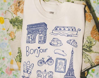 May include: Cream-colored t-shirt with a blue graphic design featuring the Eiffel Tower, Arc de Triomphe, and other French icons. The word "Bonjour" is written in blue script. The shirt is folded and resting on a floral patterned surface.