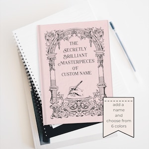 May include: A pink journal with the text "The Secretly Brilliant Masterpieces of Custom Name" in black script. The cover features an ornate arch design. A pen rests on the white surface next to the journal. A tag says "add a name and choose from 6 colors."