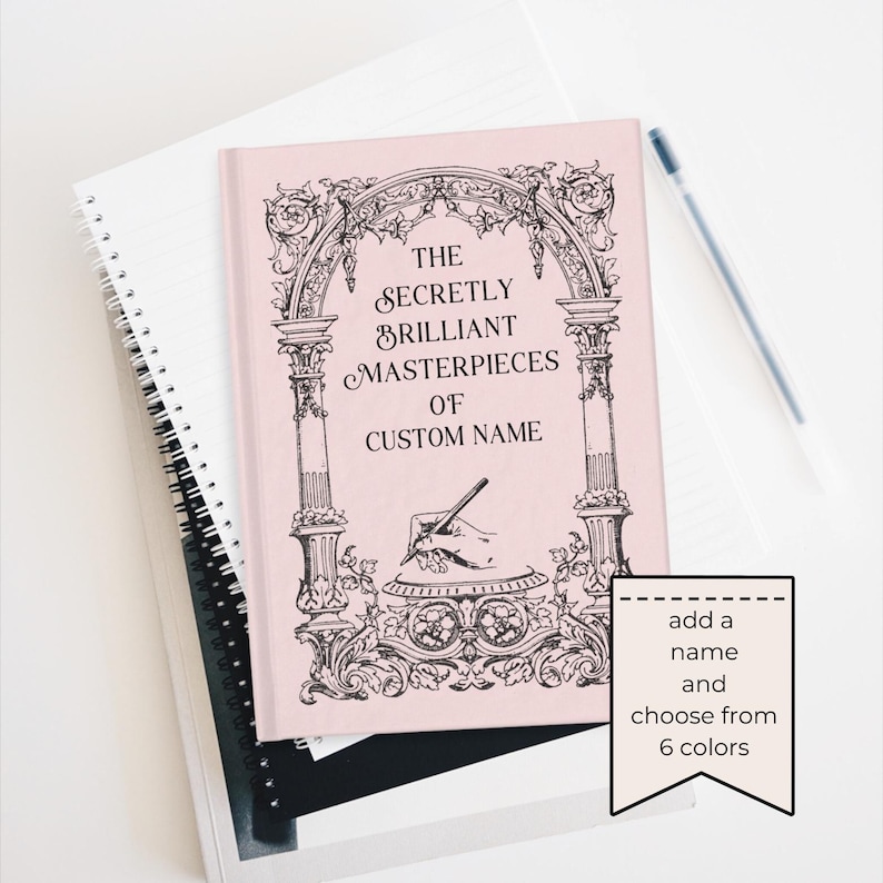 May include: A pink journal with the text "The Secretly Brilliant Masterpieces of Custom Name" in black script. The cover features an ornate arch design. A pen rests on the white surface next to the journal. A tag says "add a name and choose from 6 colors."