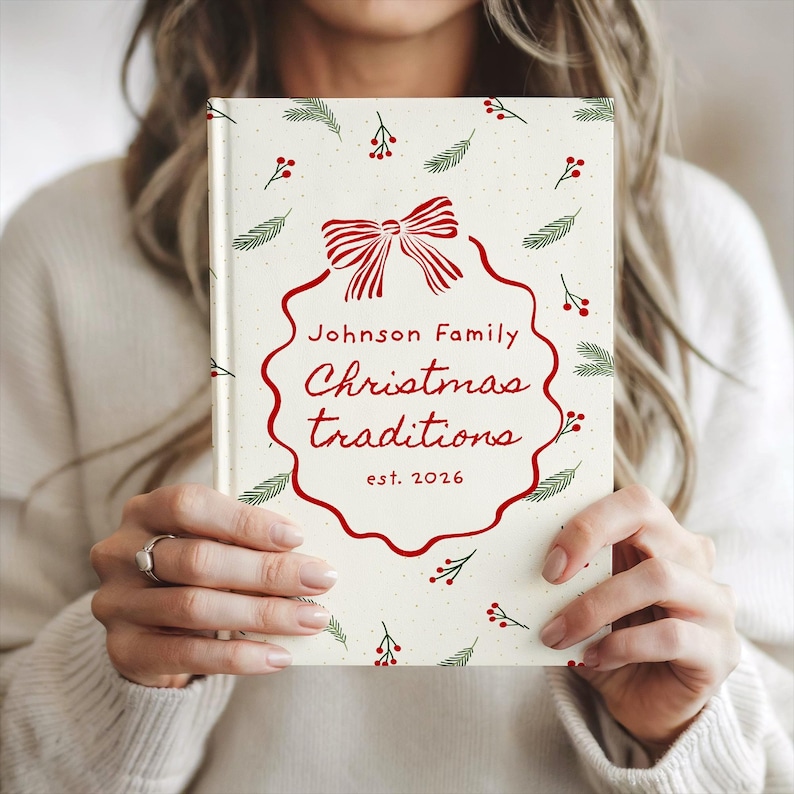 May include: A hardcover book with a cream-colored cover featuring a red scalloped frame, a red bow, and green pine sprigs with red berries. The text reads Johnson Family Christmas traditions est. 2026 in red script and block lettering.
