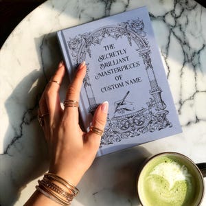 Journal shown at a -45 degree angle in the color "sky" with a woman's hand resting on the left edge of the book on top of a gray and white marble countertop. There is a filled cup with matcha tea at the bottom edge of the frame.