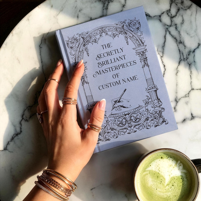 Journal shown at a -45 degree angle in the color "sky" with a woman's hand resting on the left edge of the book on top of a gray and white marble countertop. There is a filled cup with matcha tea at the bottom edge of the frame.