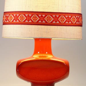 May include: A vibrant orange table lamp with a cylindrical lampshade. The lampshade is beige with a decorative band of red and orange geometric patterns. The lamp base is a glossy orange with a unique, rounded shape.