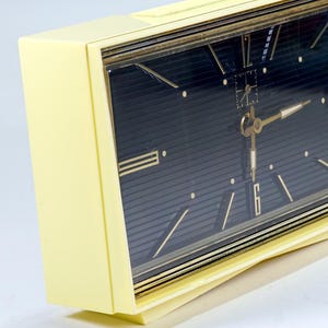 May include: A vintage, rectangular alarm clock with a pale yellow case and a dark blue face. The clock features gold-colored hands and markers, with a clear glass cover. The design is retro, with a classic aesthetic.