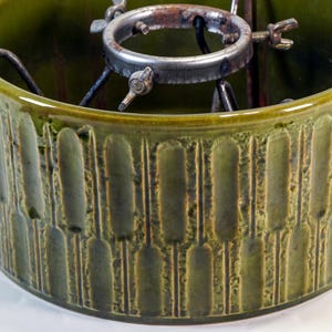 May include: A green ceramic Christmas tree stand with a textured, vertical pattern. The stand has a metal mechanism inside to hold a tree trunk. The stand is round and has a wide base.