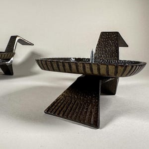 May include: Two bronze-colored metal candle holders. Each has a circular, ribbed dish for the candle and a stylized, angular base. The candle holders have a modern, geometric design. The candle holders are made of metal.