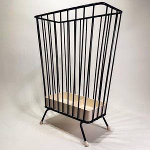 May include: A tall, rectangular storage basket constructed from thin black metal rods arranged in a vertical pattern. The basket features a cream-colored plastic base with a central divider and stands on four black metal legs with white feet.