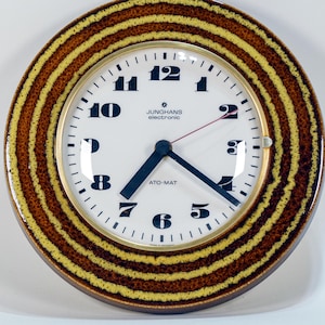 Op de afbeelding: Een ronde wandklok met een witte wijzerplaat, zwarte cijfers en blauwe wijzers. De buitenste ring van de klok heeft een bruin en geel gestreept ontwerp. De wijzerplaat leest "Junghans electronic ATO-MAT".
