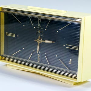 May include: A vintage, rectangular alarm clock with a cream-colored case and a dark blue face. The clock features gold-colored hands, hour markers, and a gold-colored frame. The clock's face has a textured, striped pattern.