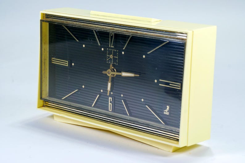 May include: A vintage, rectangular alarm clock with a cream-colored case and a dark blue face. The clock features gold-colored hands, hour markers, and a gold-colored frame. The clock's face has a textured, striped pattern.