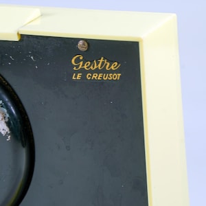 May include: Close-up of a vintage clock with a cream-colored frame and a dark green face. The clock face has a key and the text "Gestre LE CREUSOT" in gold lettering. The clock shows signs of wear.