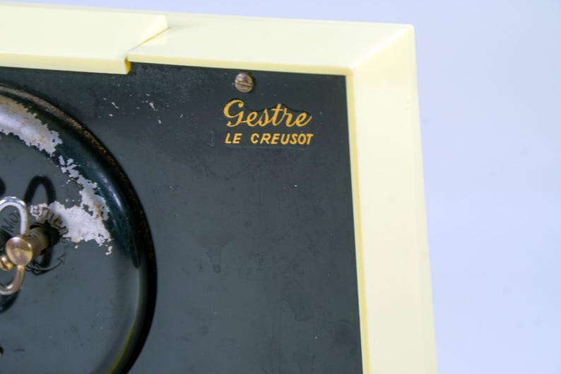 May include: Close-up of a vintage clock with a cream-colored frame and a dark green face. The clock face has a key and the text "Gestre LE CREUSOT" in gold lettering. The clock shows signs of wear.