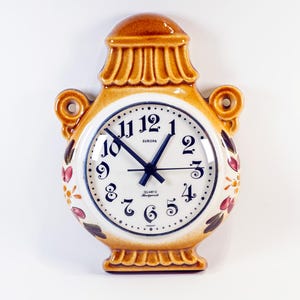 May include: A ceramic wall clock in a vase shape, featuring a white clock face with black numerals and hands. The clock has a brown and tan color scheme with floral accents. The clock reads "EUROPA" and "Quartz Made in Hong Kong".