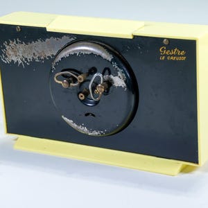 May include: A vintage, rectangular clock with a black face and a cream-colored frame. The clock face has a circular design with winding keys. The text "Gestre LE CREUSOT" is printed on the clock face.