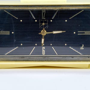 May include: A vintage cream-colored rectangular clock with a dark blue face. The clock features gold-colored hands and markers, with the numbers 12 and 6 visible. The clock has a gold-colored trim and the brand name "JAZ" is visible.