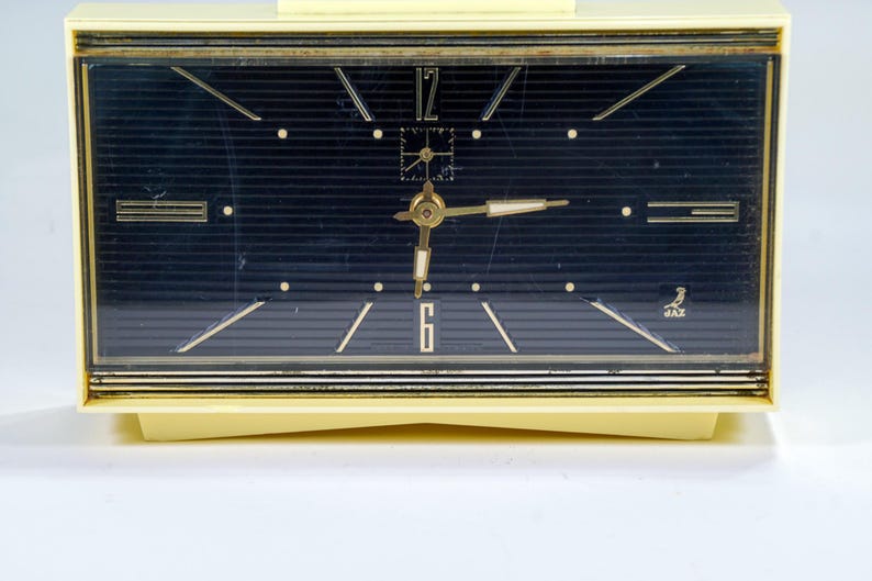 May include: A vintage cream-colored rectangular clock with a dark blue face. The clock features gold-colored hands and markers, with the numbers 12 and 6 visible. The clock has a gold-colored trim and the brand name "JAZ" is visible.