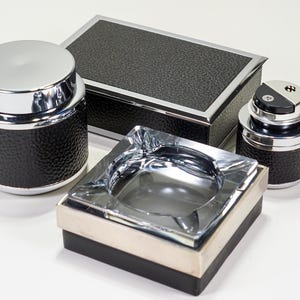 Pode incluir: Uma coleção de acessórios pretos e prateados, incluindo um recipiente cilíndrico com tampa, uma caixa retangular, um cinzeiro quadrado e um isqueiro. Os itens têm uma superfície preta texturizada e detalhes prateados brilhantes.