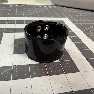May include: A glossy black cuff bracelet with a buckle closure. The bracelet has a wide band and is secured with metal rivets. The surface reflects light, highlighting its smooth texture. This accessory is designed to be worn around the wrist.