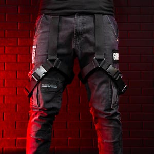 May include: Dark gray denim cargo pants with black straps and buckles. The pants have multiple pockets and patches with text. The background is a red brick wall.