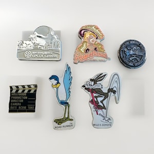 May include: A collection of enamel pins featuring various designs. Included are pins with the text "Walt Disney World EPCOT Center", "Rosie O'Grady", a film clapperboard, the Road Runner, Wile E. Coyote, and "Babyland General Hospital".