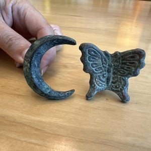 May include: Two antique bronze decorative pieces: a crescent moon and a butterfly. The moon is curved, with a dark, aged patina. The butterfly has detailed wing patterns and stands on small feet. Both are on a wooden surface.
