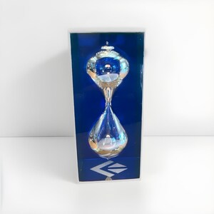 May include: A clear glass hourglass with a silver-toned metallic finish on the top and bottom bulbs, encased in a rectangular blue box. The text Rite Lite is visible on the top bulb, and a geometric logo is printed at the bottom of the box.