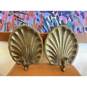 May include: A pair of brass wall sconces, each featuring a large, fan-shaped shell backplate with vertical ridges and a single cylindrical candle holder attached to the lower center. The metal has a polished, warm golden finish throughout.