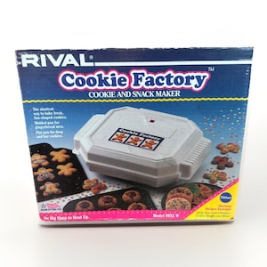 May include: A vintage Rival Cookie Factory cookie and snack maker in its original box. The box is blue with the words "Cookie Factory" in pink and white. The machine is white and has a gingerbread man design.