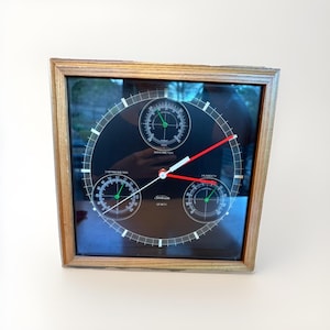 May include: A square, wooden-framed weather station with a black face. It features a thermometer, barometer, and humidity gauge, with white markings and green needles. The clock hands are white and red. The brand name "Sunbeam Quartz" is printed on the face.