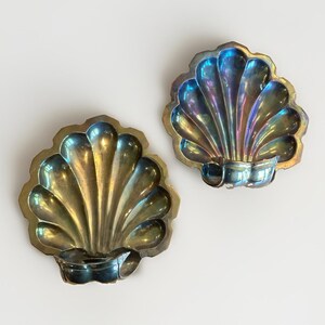 May include: Two metallic shell-shaped dishes with a scalloped edge and a reflective, iridescent finish. Each dish features a curved, fluted design radiating from a central base, displaying shifting tones of gold, blue, and purple across the surface.