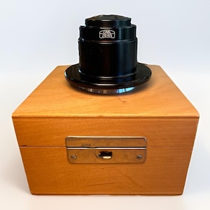 May include: A black Carl Zeiss camera lens resting on top of a light brown wooden box. The box features a rectangular metal latch centered on the front panel. The lens has a cylindrical body with a wide base and the brand name printed in white.