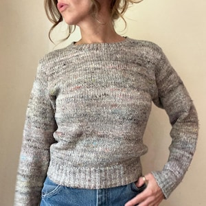 May include: A gray knit sweater with a crew neck and long sleeves. The sweater has flecks of color throughout and a ribbed hem. The person is wearing blue jeans.