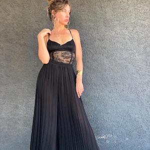 May include: Black jumpsuit with a lace bodice and pleated skirt. The top features spaghetti straps and a sweetheart neckline. The skirt is floor-length and flows from the waist. The outfit is paired with black boots and gold jewelry.