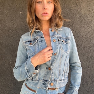 May include: Light blue denim jacket with button closures and chest pockets. The jacket is paired with matching light blue jeans. The outfit features brown leather accents and a classic, casual style.
