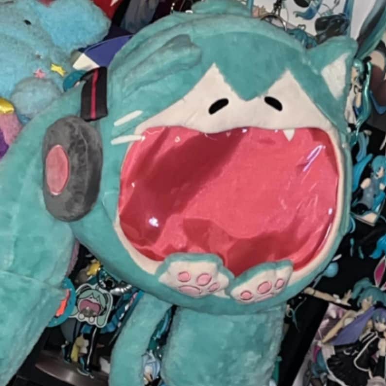 May include: A large, plush teal cat-like creature with a wide-open mouth revealing a pink interior. The plush has black eyes, white fangs, and a black and red circular detail on the side. The plush is surrounded by other plush toys and anime-style merchandise.