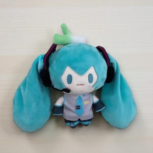 May include: A plush doll featuring a cartoon character with turquoise hair in long pigtails, wearing a gray vest, blue tie, and headset. The doll has a small green sprout on its head and is designed in a chibi style.