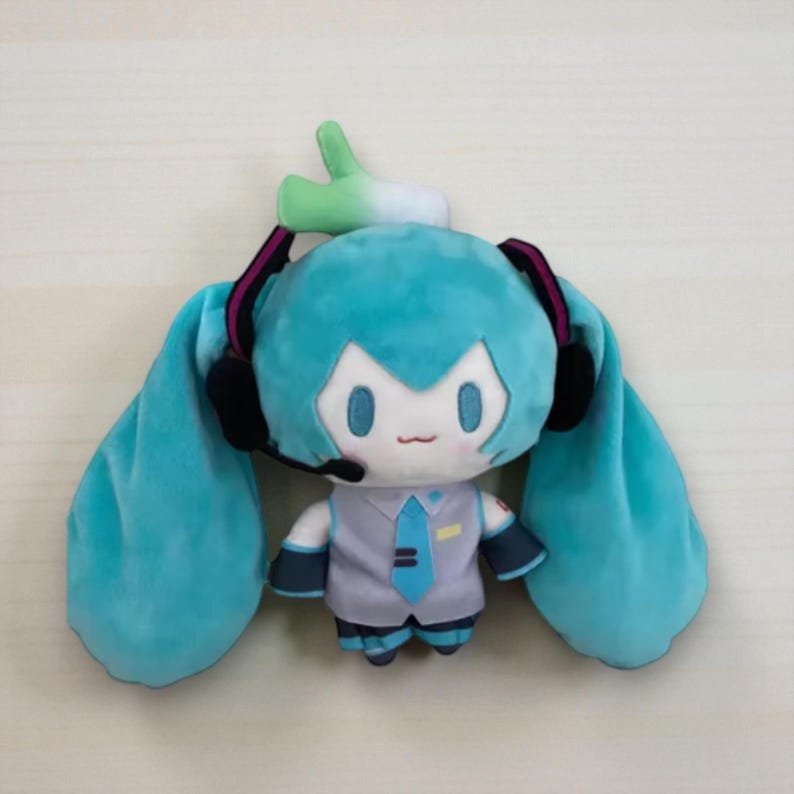 May include: A plush doll featuring a cartoon character with turquoise hair in long pigtails, wearing a gray vest, blue tie, and headset. The doll has a small green sprout on its head and is designed in a chibi style.