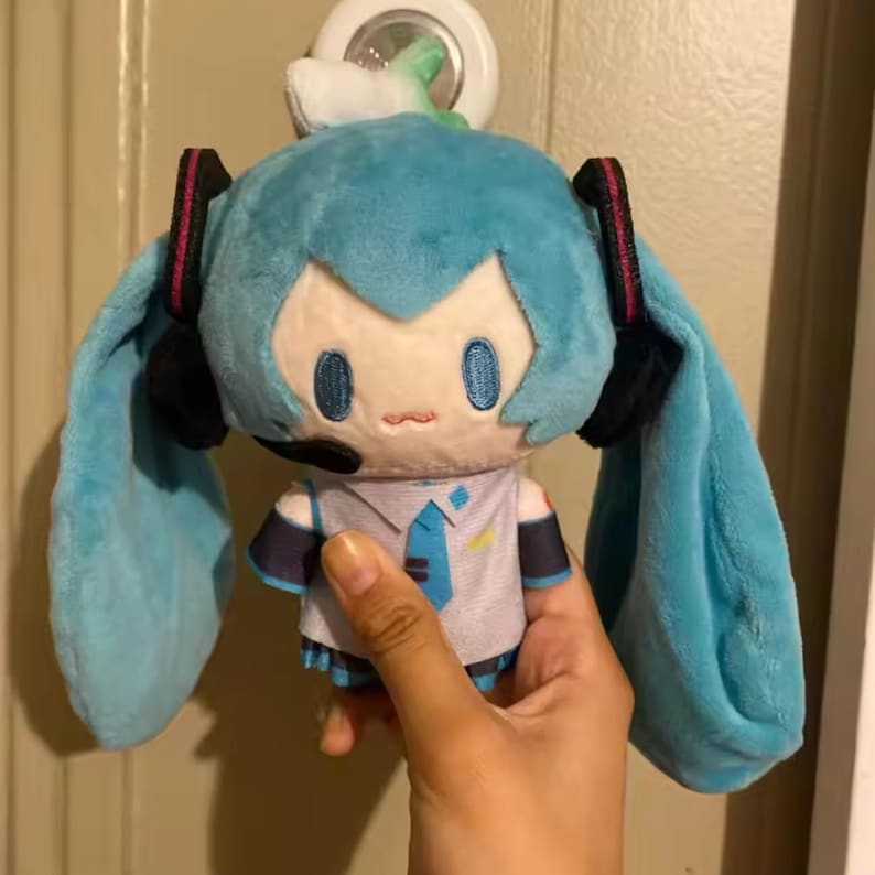May include: A plush doll with light blue hair and long pigtails, wearing a white shirt with a blue tie and black headphones. The doll has a small mouth and blue eyes. The doll is being held by a hand.