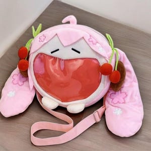 Puede incluir: Un bolso de hombro de felpa rosa con un diseño de cara de dibujos animados. El bolso presenta una gran boca roja, ojos cerrados y detalles de cereza. Tiene una correa rosa y está decorado con formas de flores.