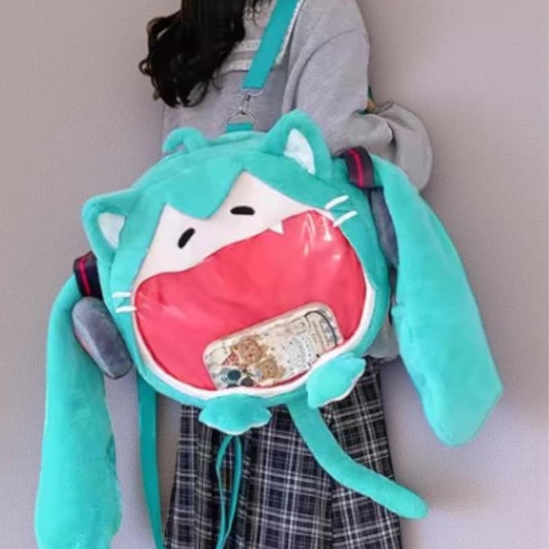 May include: A teal plush backpack shaped like a cartoon character's face with large, open mouth. The backpack has long, floppy ears and a clear pocket displaying a phone. The straps are teal and the backpack has a tail.