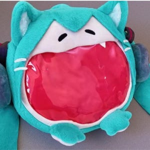 May include: A teal plush backpack shaped like a cartoon cat's head, featuring white accents, black eyes, and a large, open red mouth. The backpack has cat ears and paws, with a clear, plastic-like opening for storage.